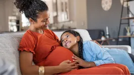 Smiling photo of black pregnant mother and daughter on sofa gazing lovingly at each other as they touch her tummy bump