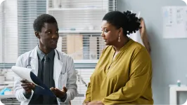 Male doctor in white coat holds up clipboard during a sitting consultation with female patient